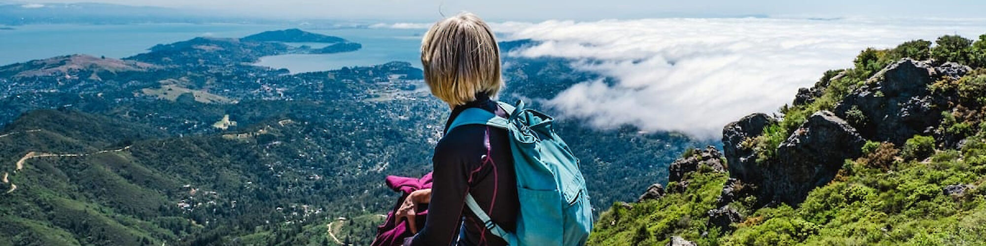 A person with a blue backpack sits on a rocky ledge, looking out over a scenic landscape with rolling hills, a distant body of water, and clouds.