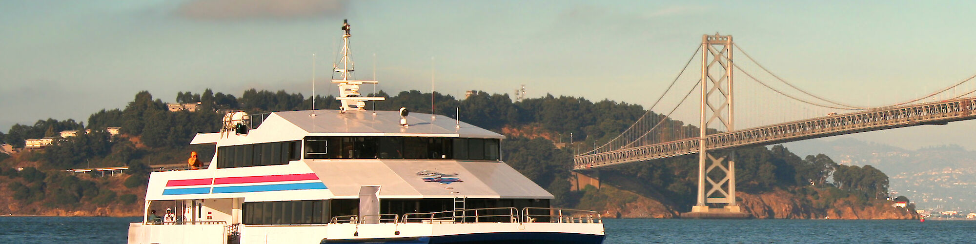 A ferry is sailing on the water with a suspension bridge and hilly landscape in the background. Bright skies and scattered clouds are present.