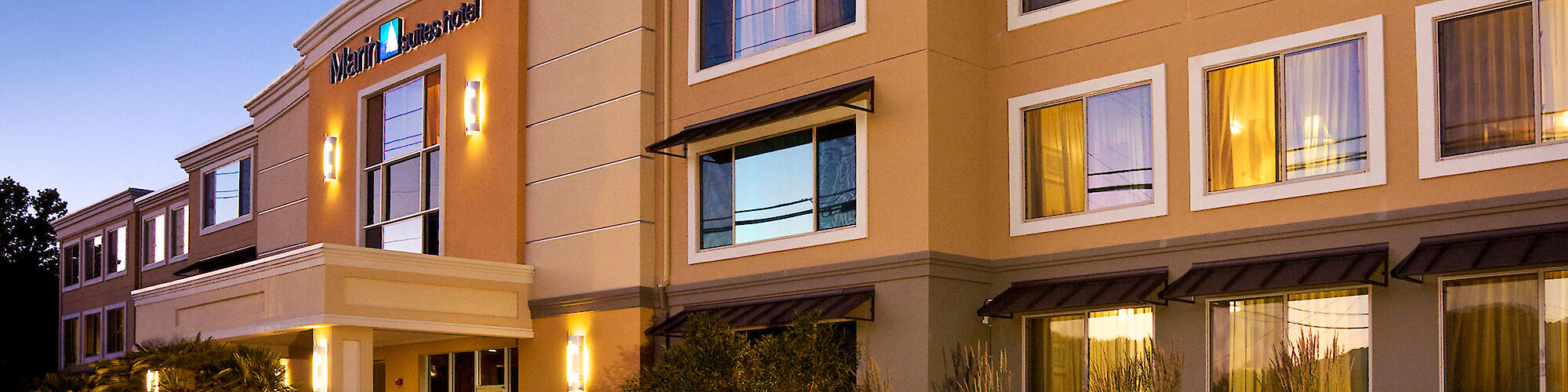 This image shows a well-lit hotel building with multiple windows and a prominently displayed entrance, surrounded by landscaped greenery.