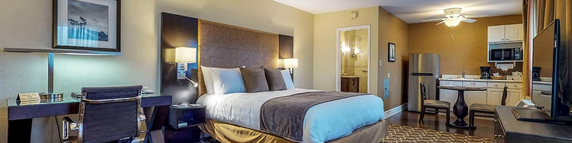 A hotel room with a bed, desk, chair, lamps, kitchenette, and TV, all in a modern and spacious layout.
