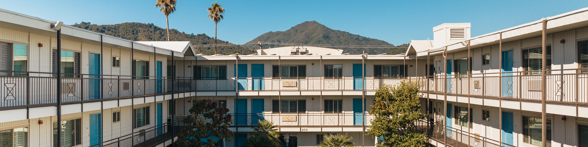 The image shows a courtyard of a hotel or apartment complex with a central swimming pool, surrounded by lounge chairs and buildings, under a clear blue sky.