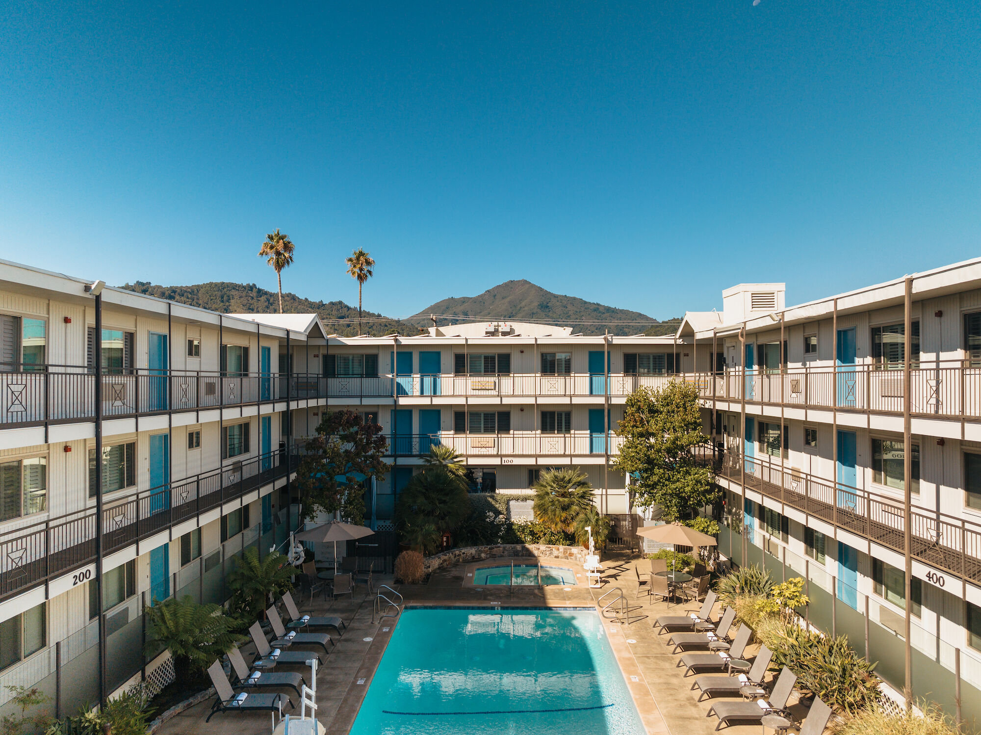 A courtyard view of a multi-story hotel with a central swimming pool surrounded by lounge chairs, umbrella-shaded tables, and distant mountains.