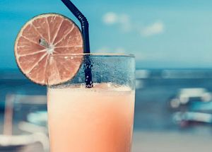 A hand holds a glass with a refreshing drink garnished with a lime slice and a straw, against a background of a beach and ocean scenery.
