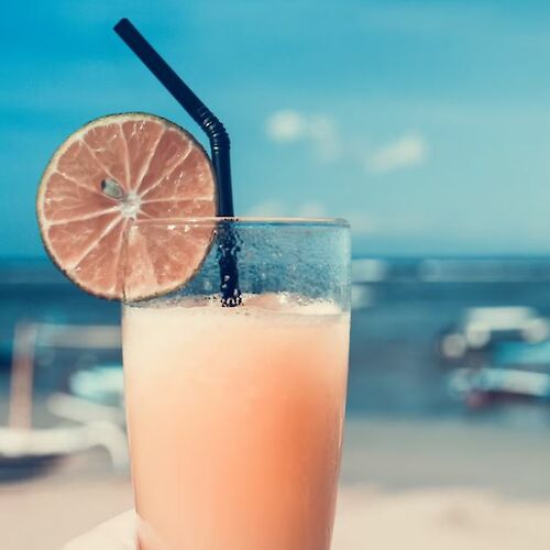 A hand holds a glass with a refreshing drink garnished with a lime slice and a straw, against a background of a beach and ocean scenery.